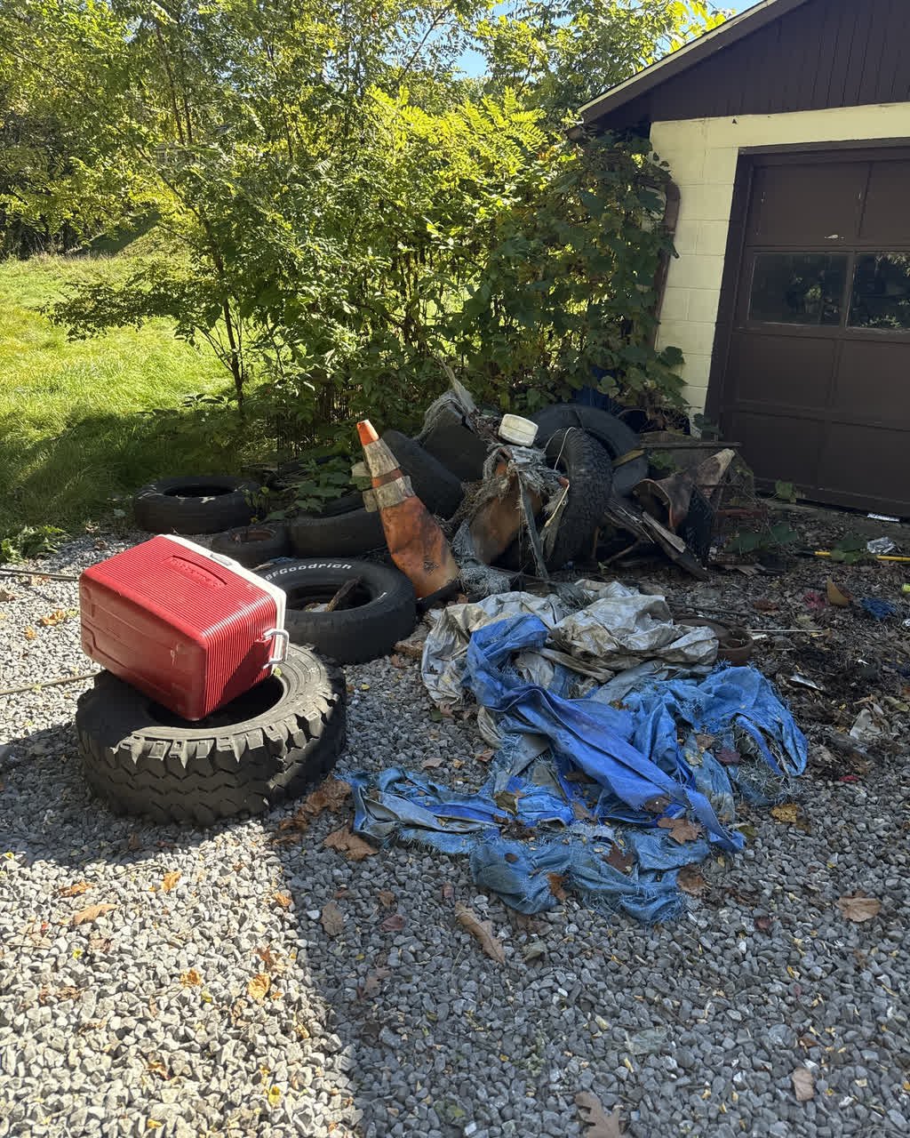 Tires, a torn blue tarp, a traffic cone, a cooler and scrap metal piled on the gravel beside a garage door